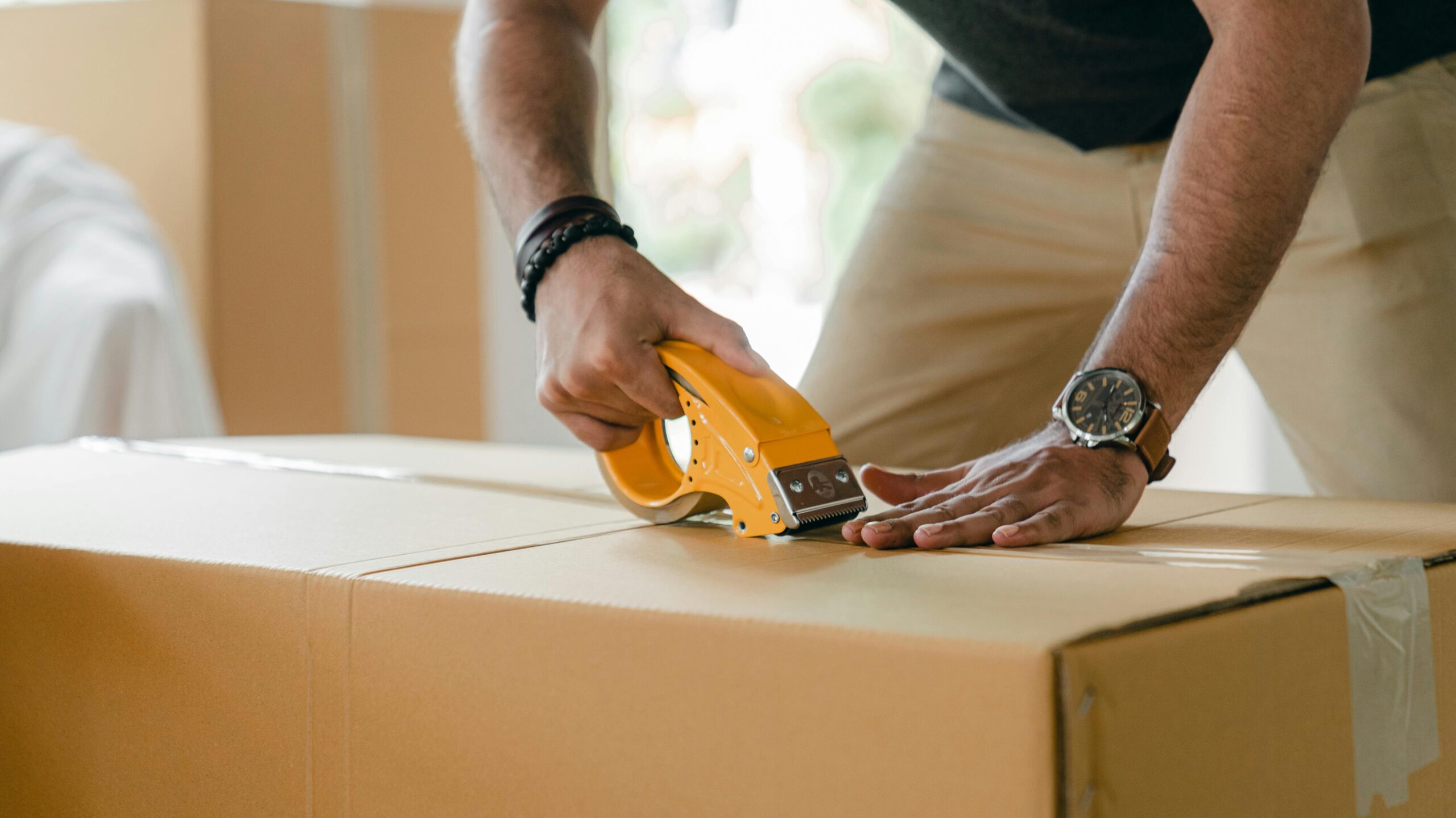 Crop unrecognizable young male in casual clothes packing cardboard box and sealing with scotch tape